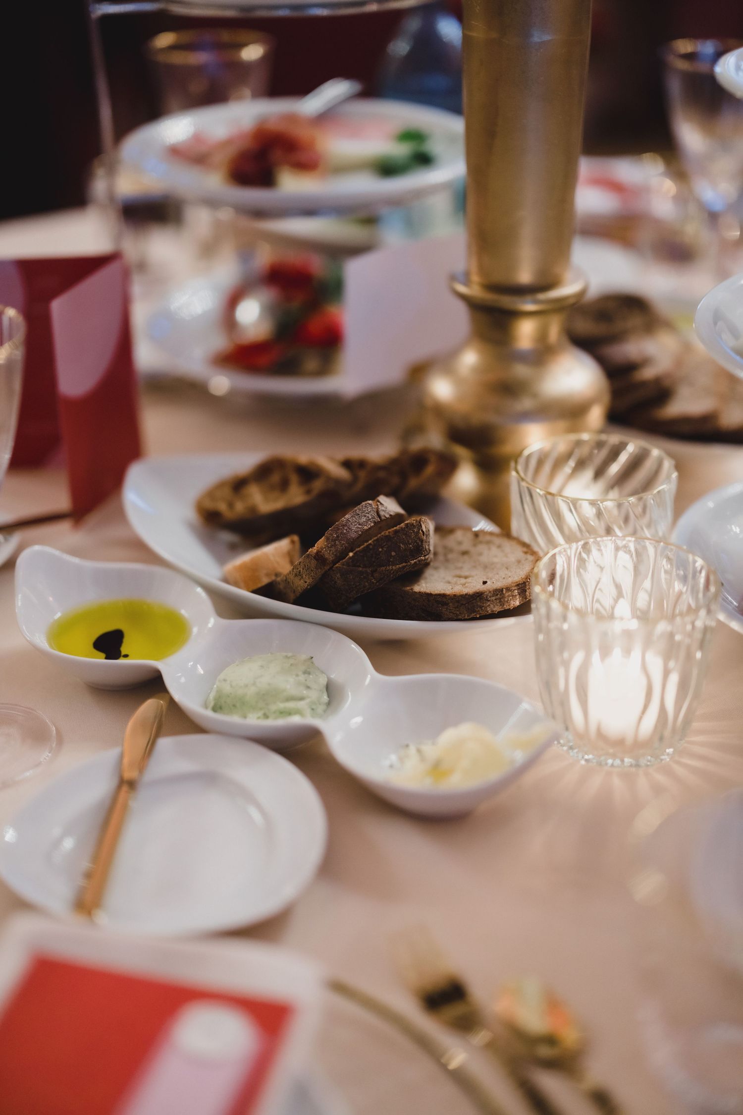 An elegant table setting with brown bread, small dishes of condiments, and a golden candlestick holder on a white tablecloth.