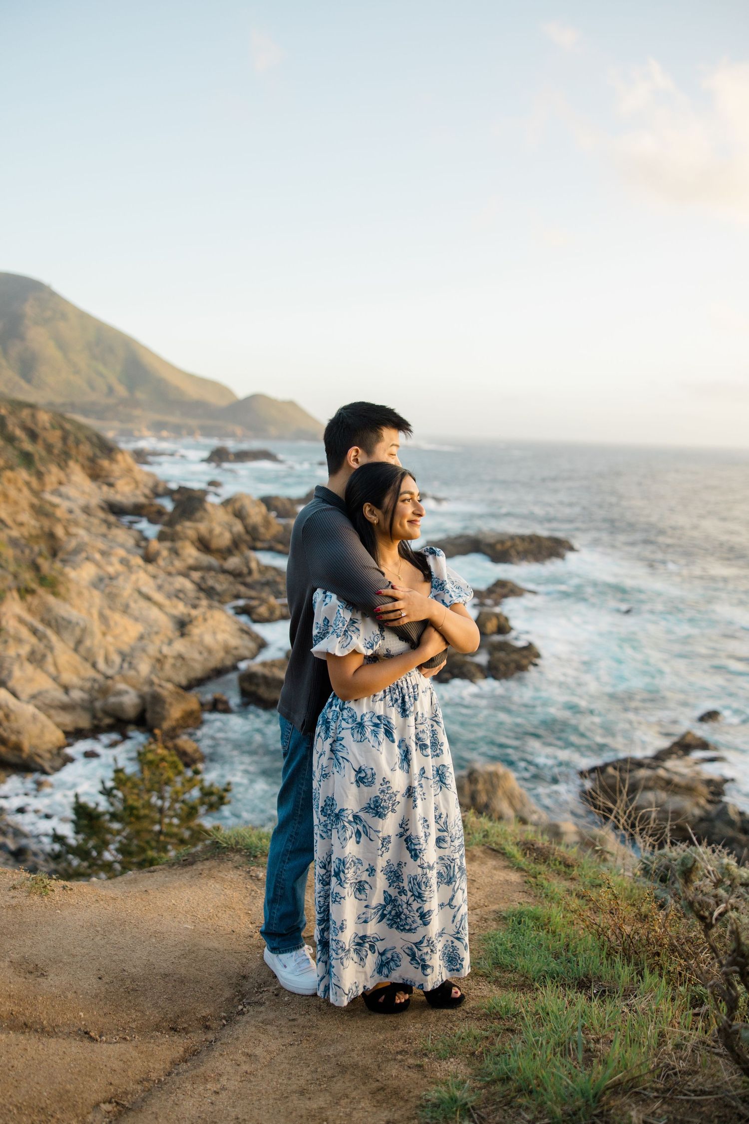 Couple embraces while standing on coastal cliff overlooking rocky Pacific shore during golden hour at Big Sur.