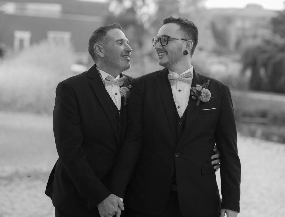 Two people in formal black suits share a joyful candid moment together during a wedding celebration.