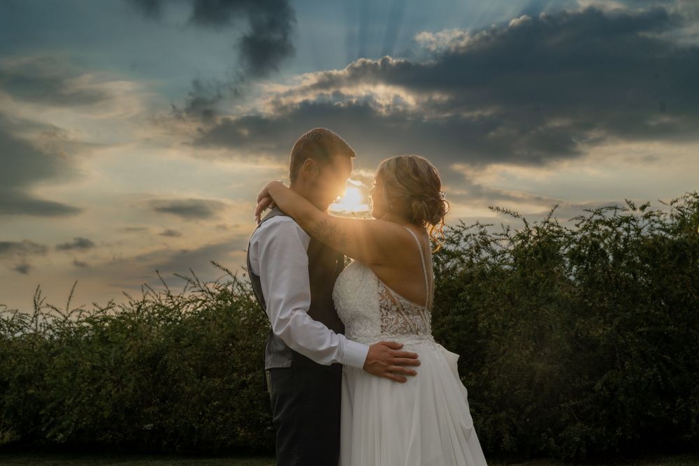 A couple embraces in a romantic sunset moment against a dramatic cloudy sky and natural landscape.
