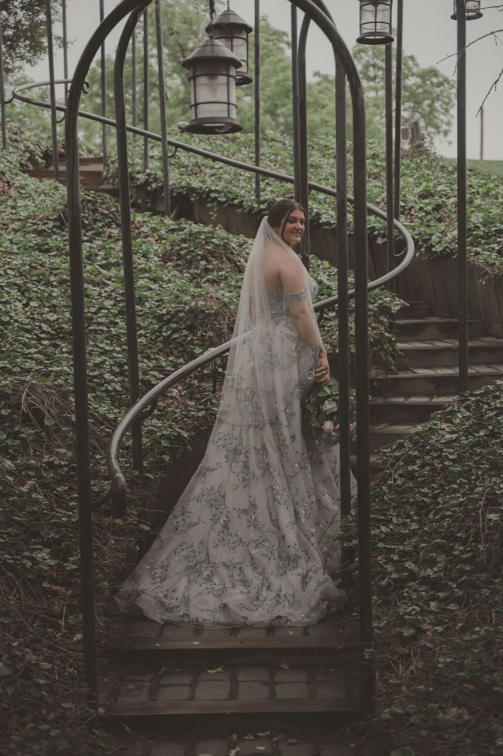 Bride in flowing gown descending outdoor spiral staircase surrounded by lush greenery.