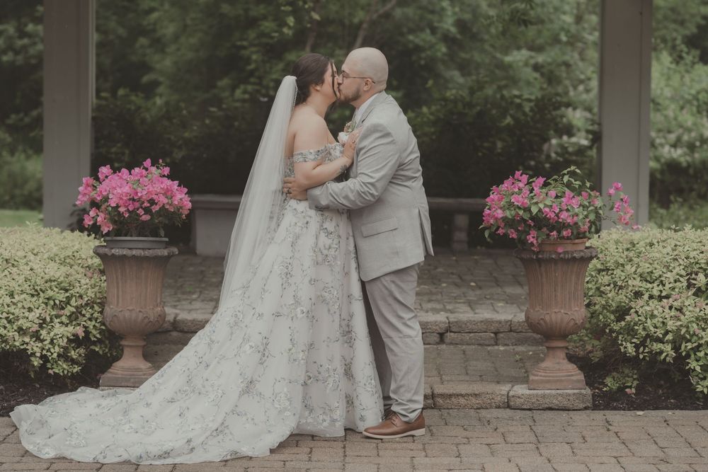 A romantic wedding couple embraces on stone steps surrounded by pink flowers and greenery in an outdoor garden setting.