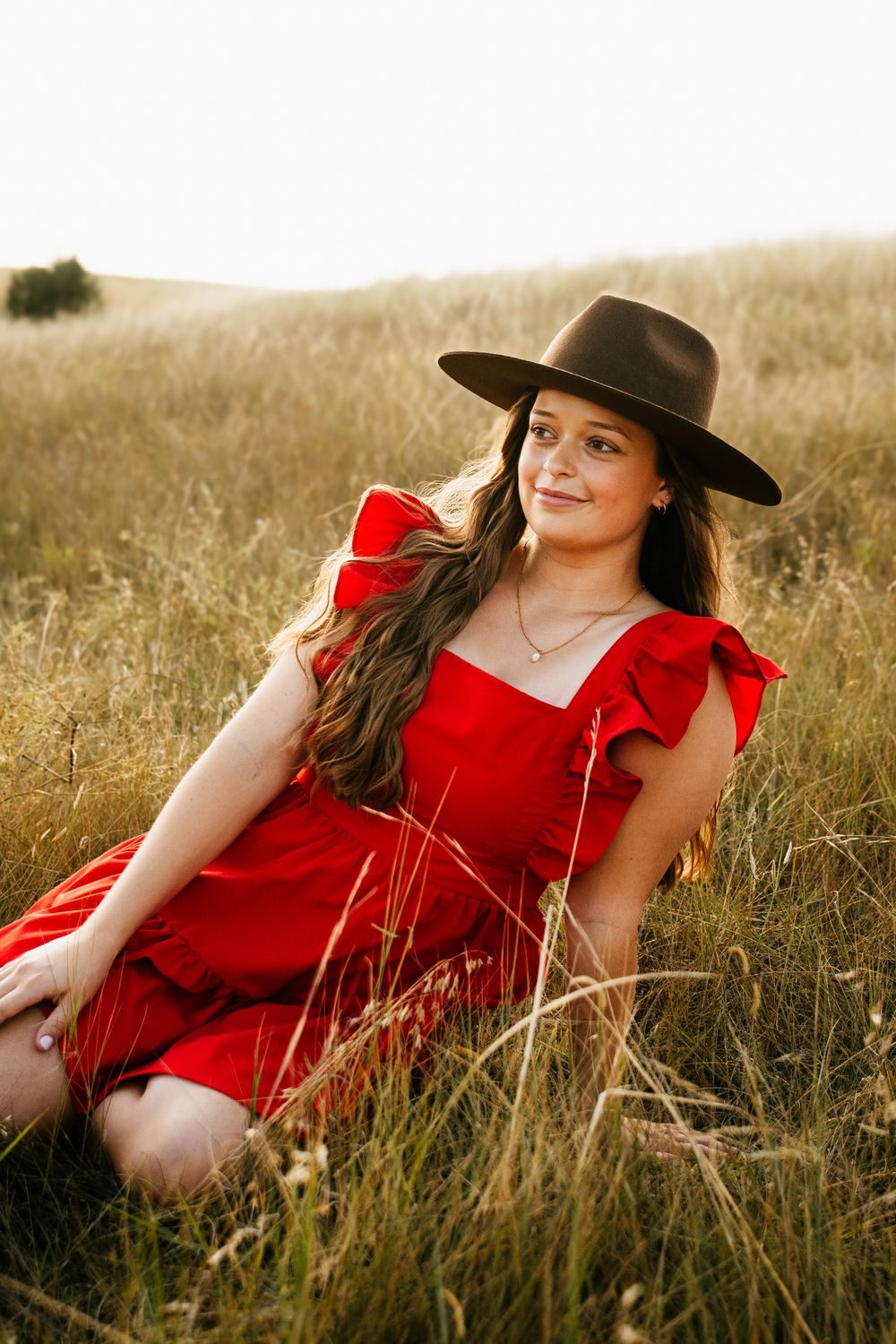A western-styled portrait of someone in a red dress sitting in tall prairie grass.