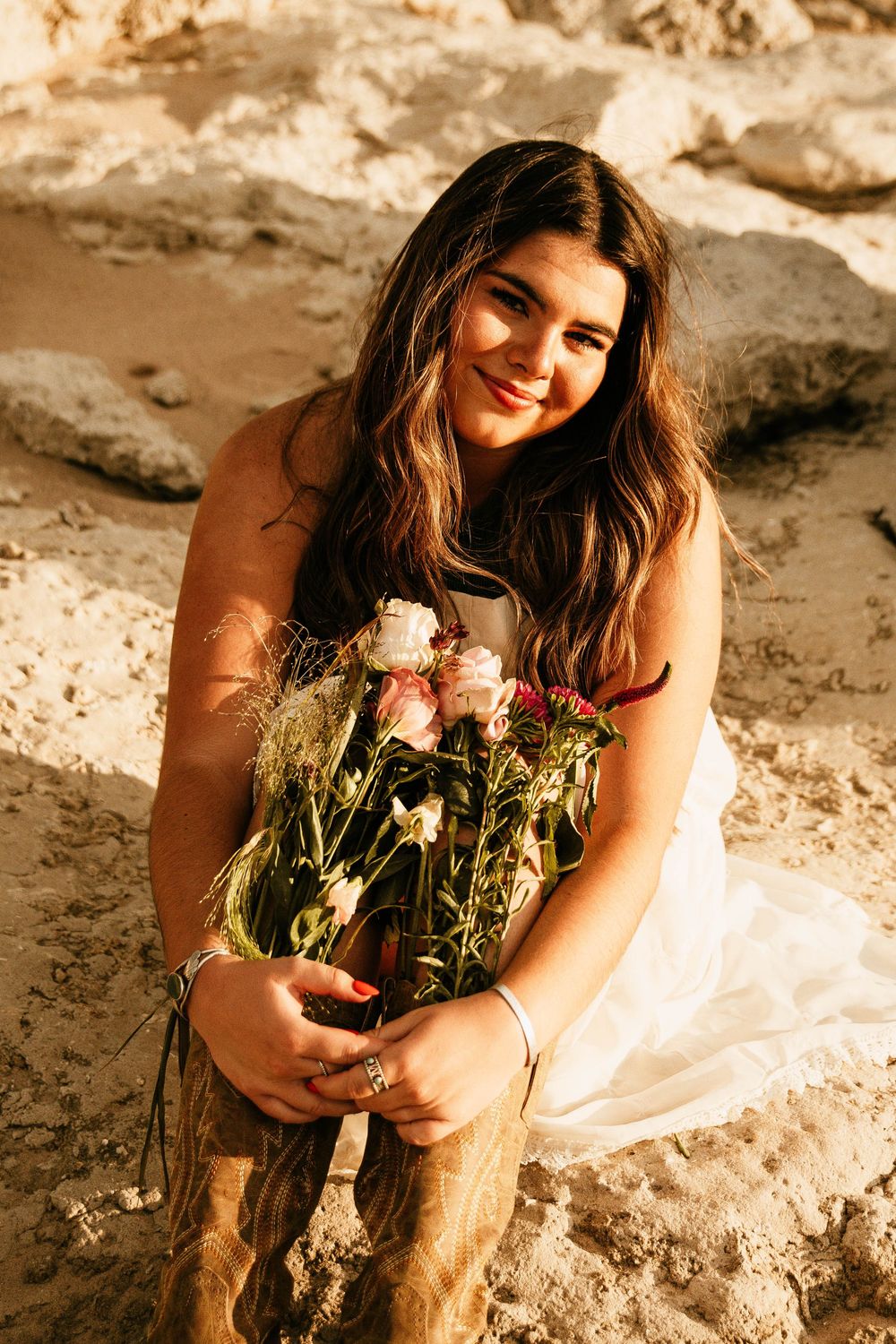 A woman in a floral dress lies on a sandy beach holding wildflowers during a golden hour photoshoot.