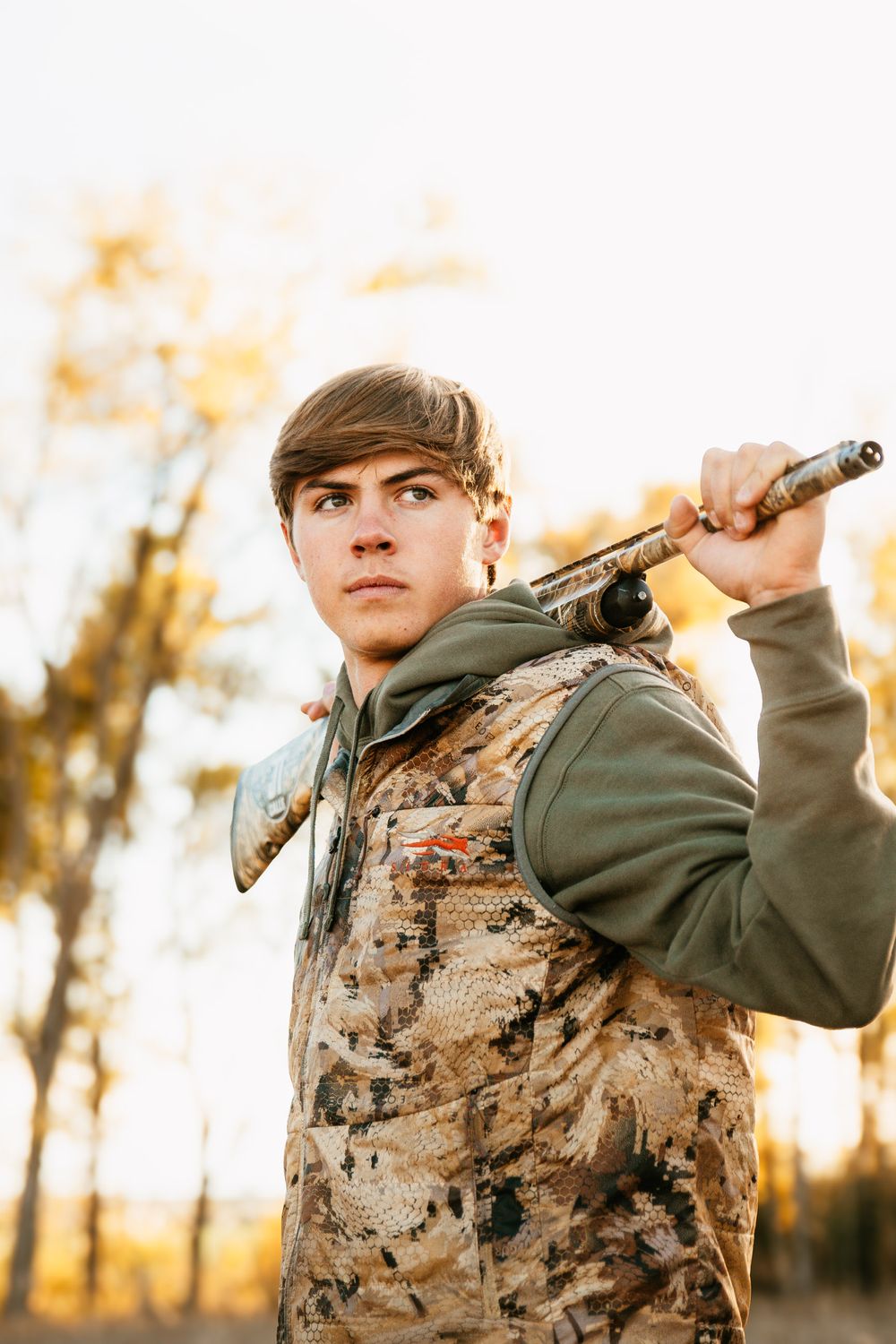 A hunter poses with a shotgun at sunset wearing camouflage gear.