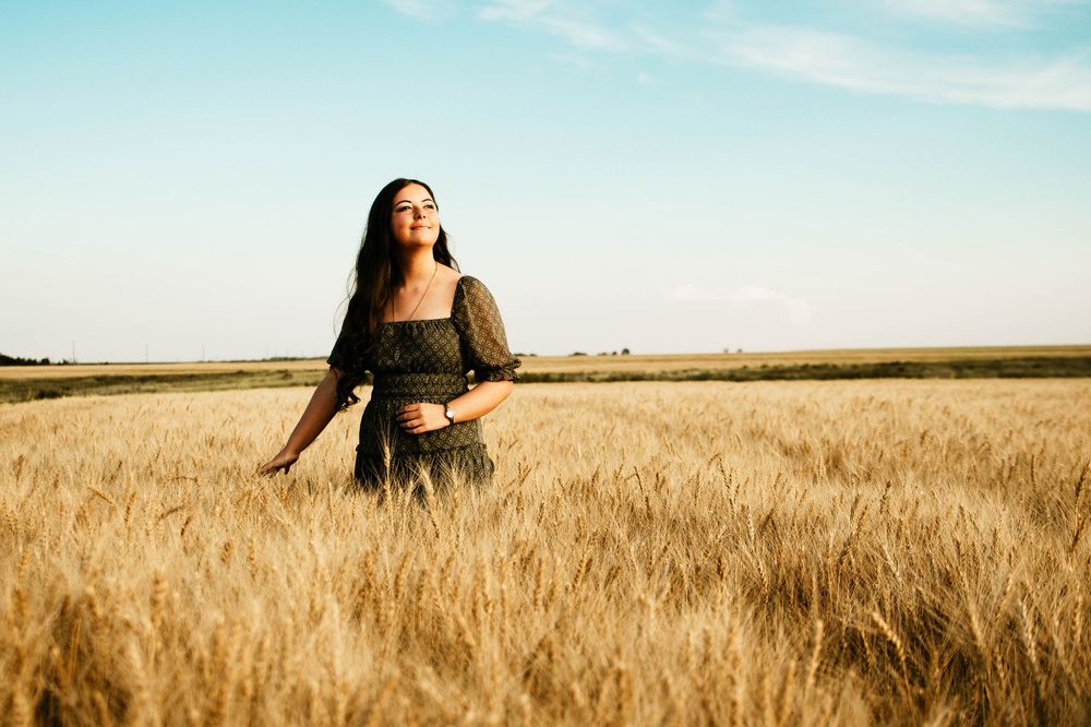 Woman in black dress walking through golden wheat field at sunset with dramatic sky in background.