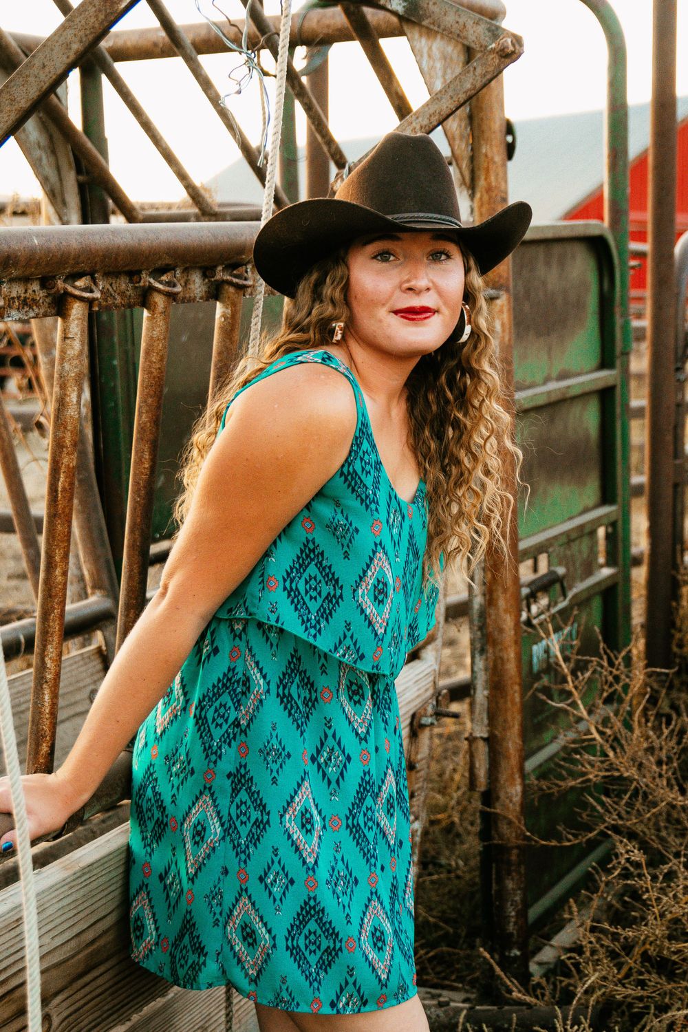 A cowgirl in a turquoise dress and black hat poses by rustic wooden beams.