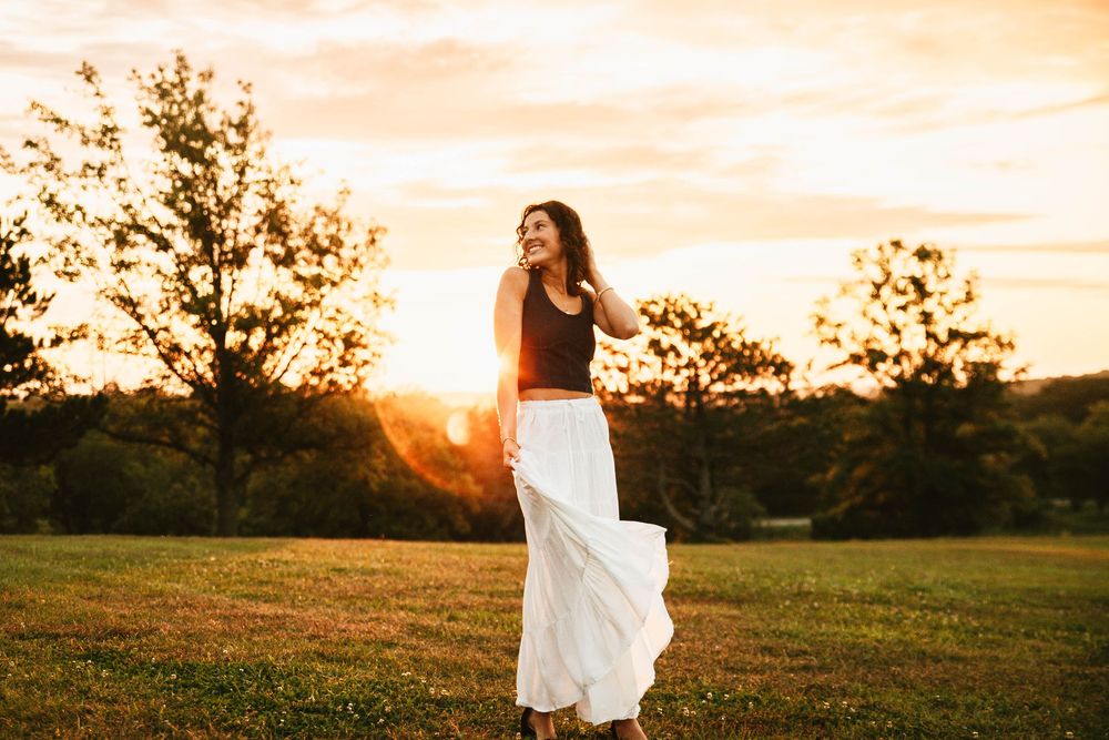 A silhouetted figure in a flowing white skirt stands in a grassy field at sunset with trees in the background.