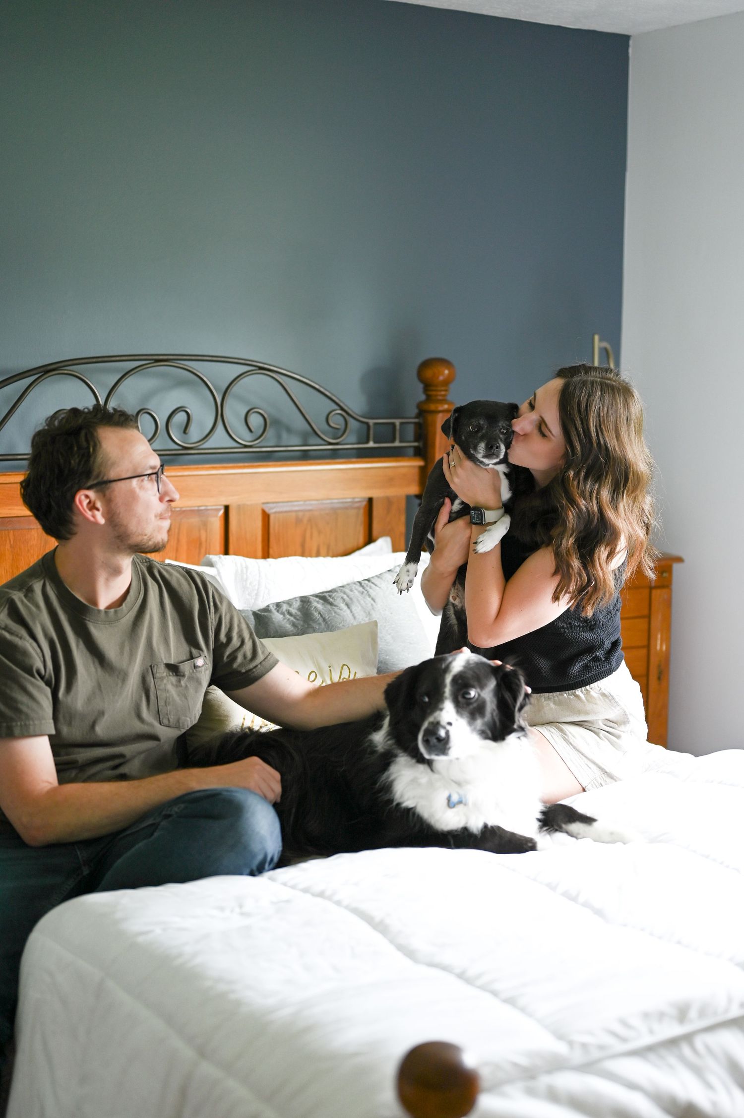 A couple sits on their bed playing with two black and white border collie dogs.
