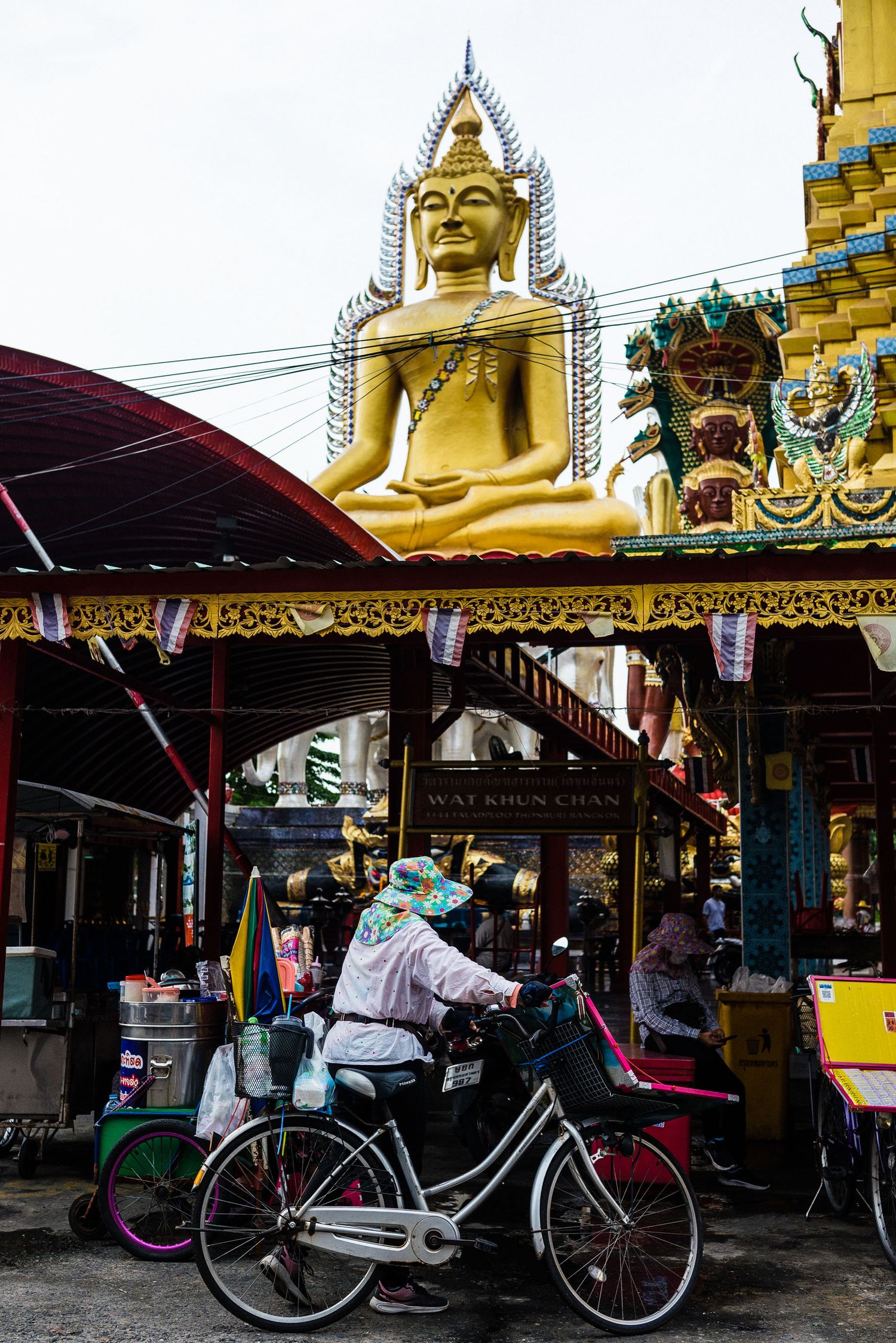 Large golden Buddha statue overlooks a street market with a cyclist passing underneath in Thailand.