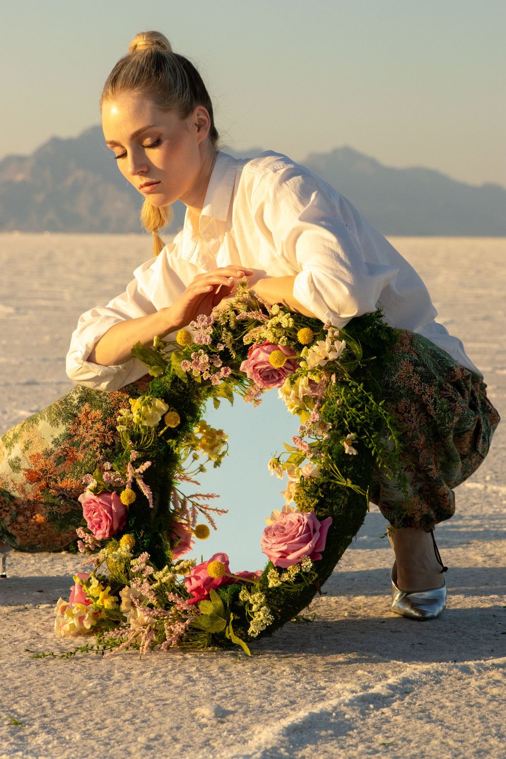 A creative portrait of a woman crouching down while holding a mirror and looking down at it with the Salt Flats in the background.