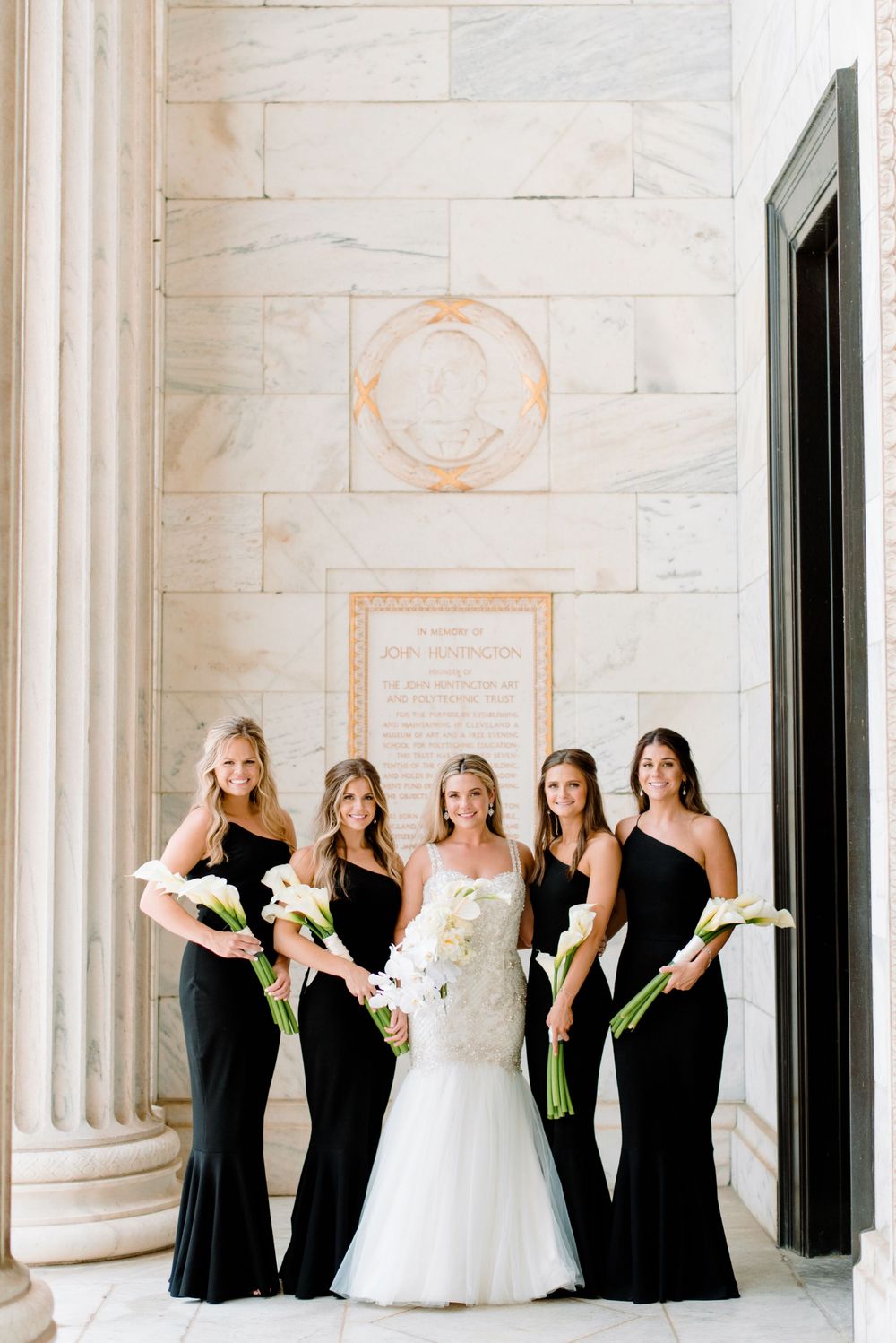 Bridal party in black dresses posing against white marble columns.