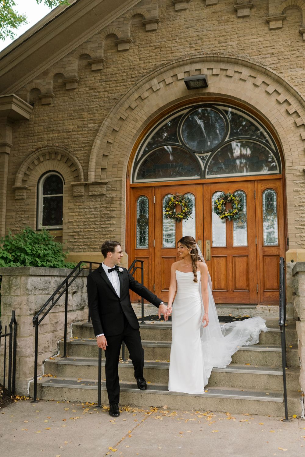 Wedding couple poses playfully in front of historic brick church with arched doorways.