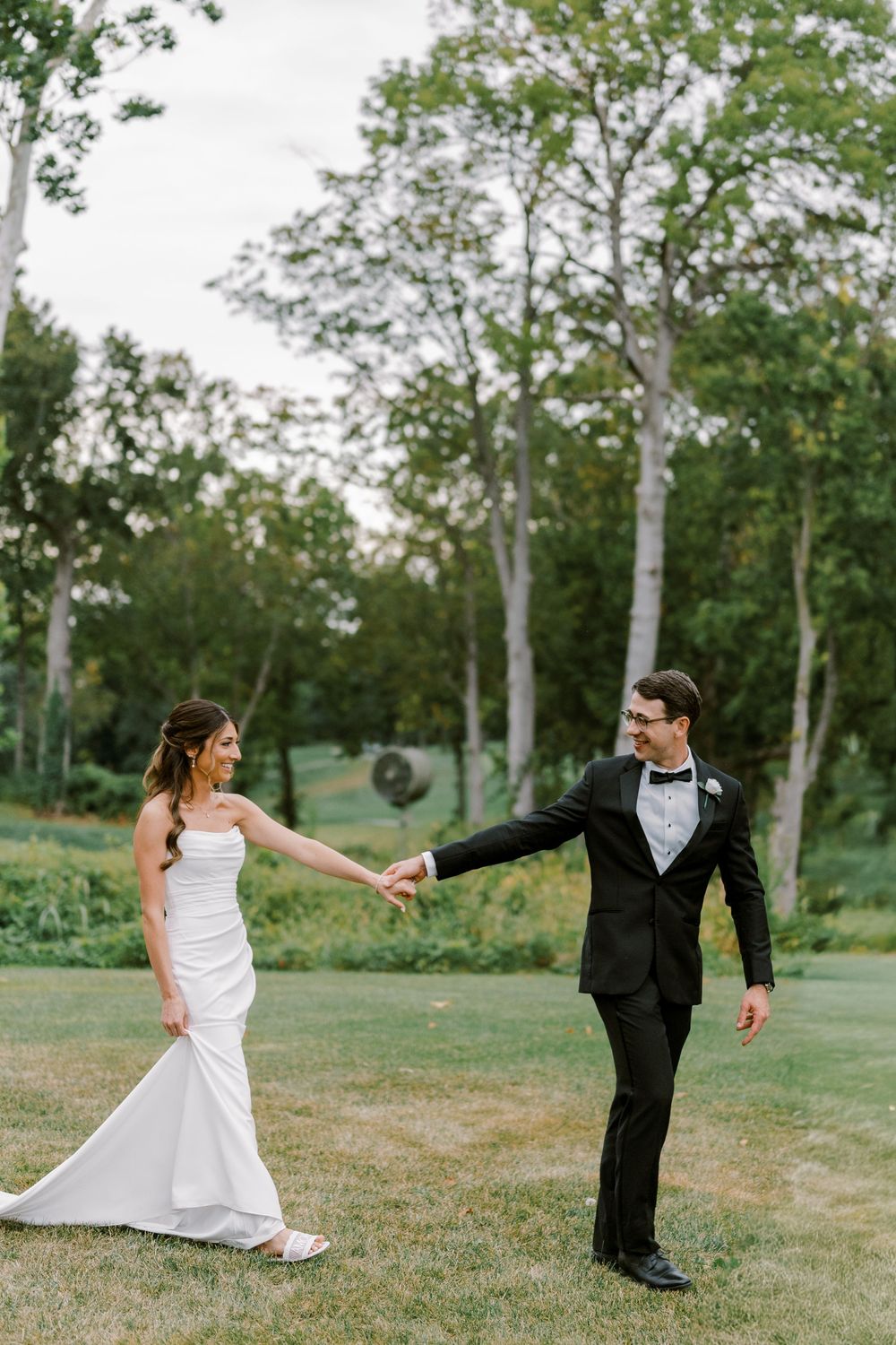 Playful wedding couple dances and spins on grassy lawn with birch trees in background.