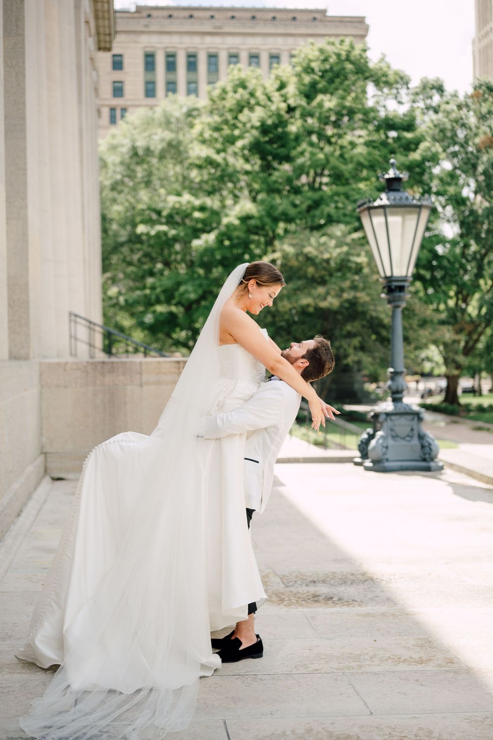 Groom dips bride in white gown and veil outside a classic building with green trees and a vintage lamp post.