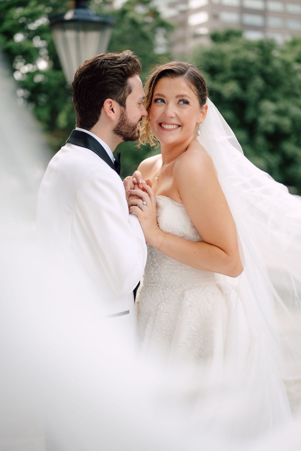 A groom in a white tuxedo kisses his smiling bride in a lace gown and veil outdoors.
