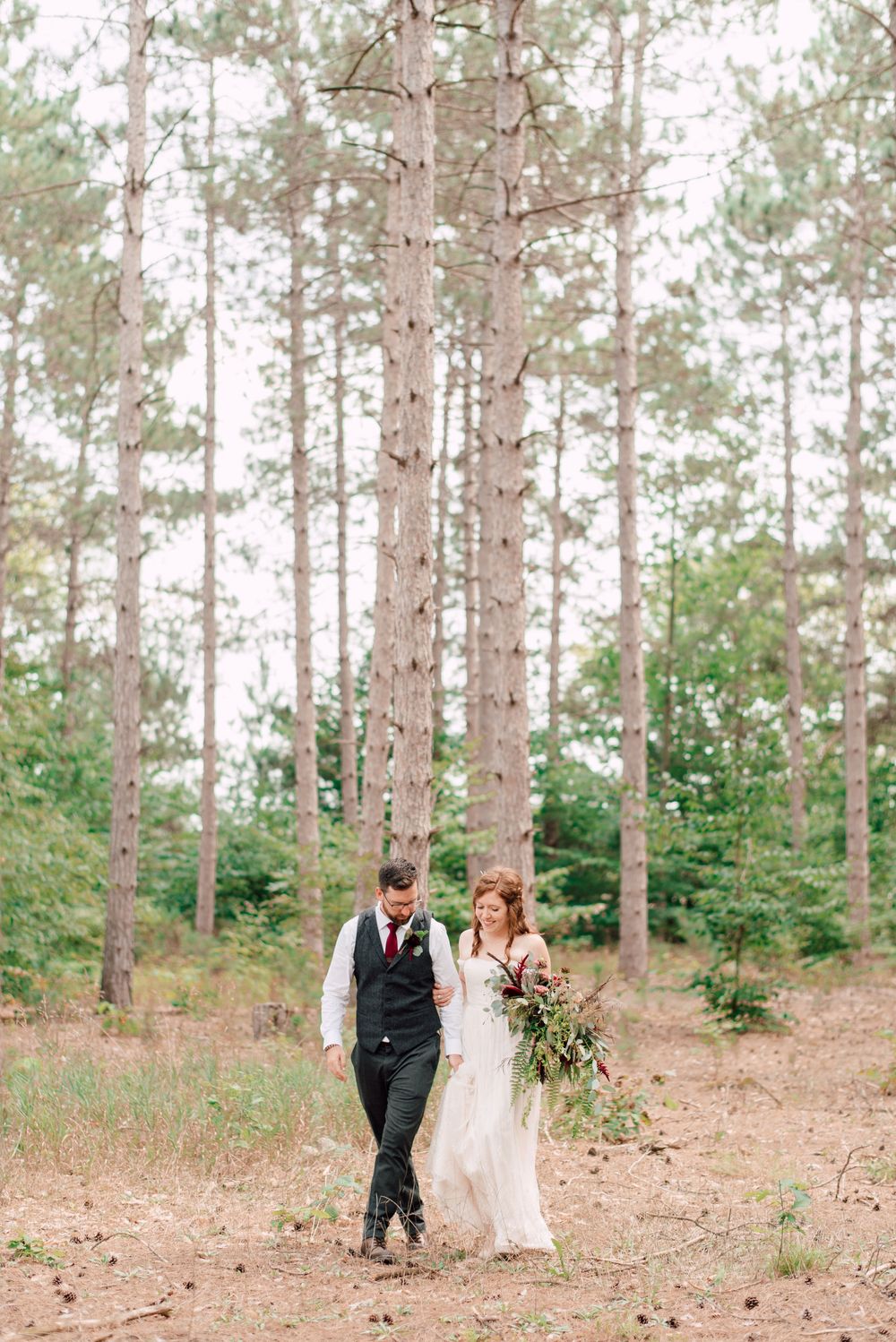 Bride and groom walk hand-in-hand through a tall pine forest, bride holding a lush burgundy bouquet.