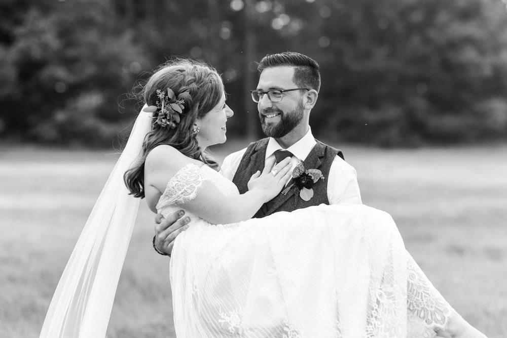 Groom carries bride in wedding dress and veil, sharing a joyful moment outdoors in black and white photo.