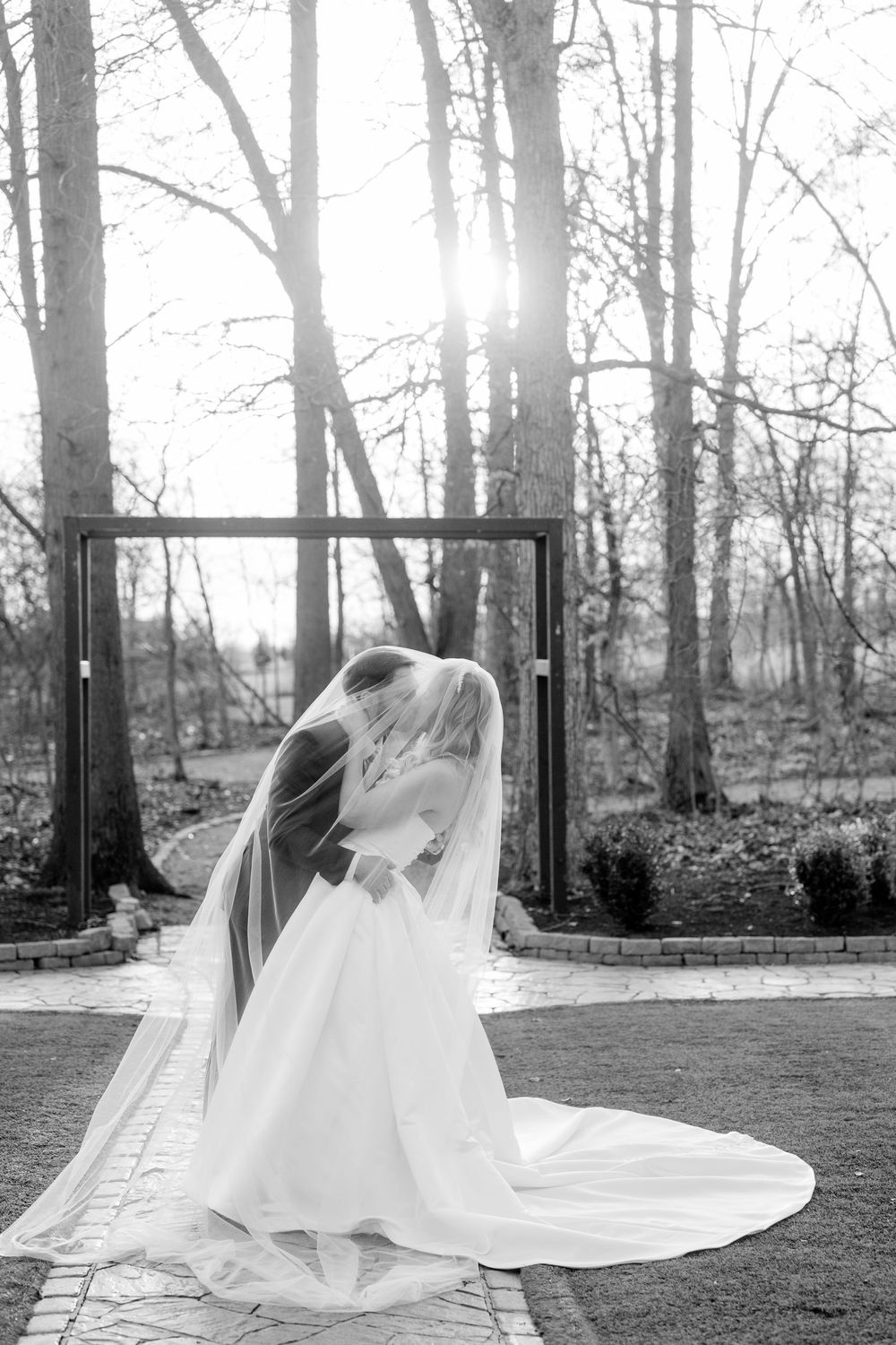 Black and white photo of a bride in a flowing gown and veil, standing outdoors near trees with sunlight behind her.