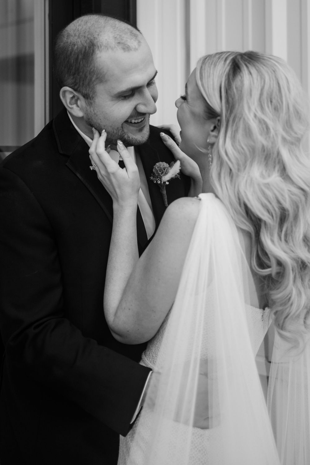 Black and white wedding photo of smiling groom and blonde bride in veil sharing a tender moment.