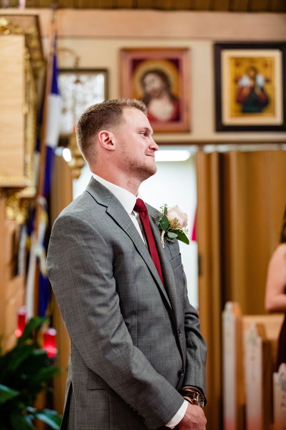 Groom in gray suit with red tie and boutonniere standing at church altar during wedding ceremony.