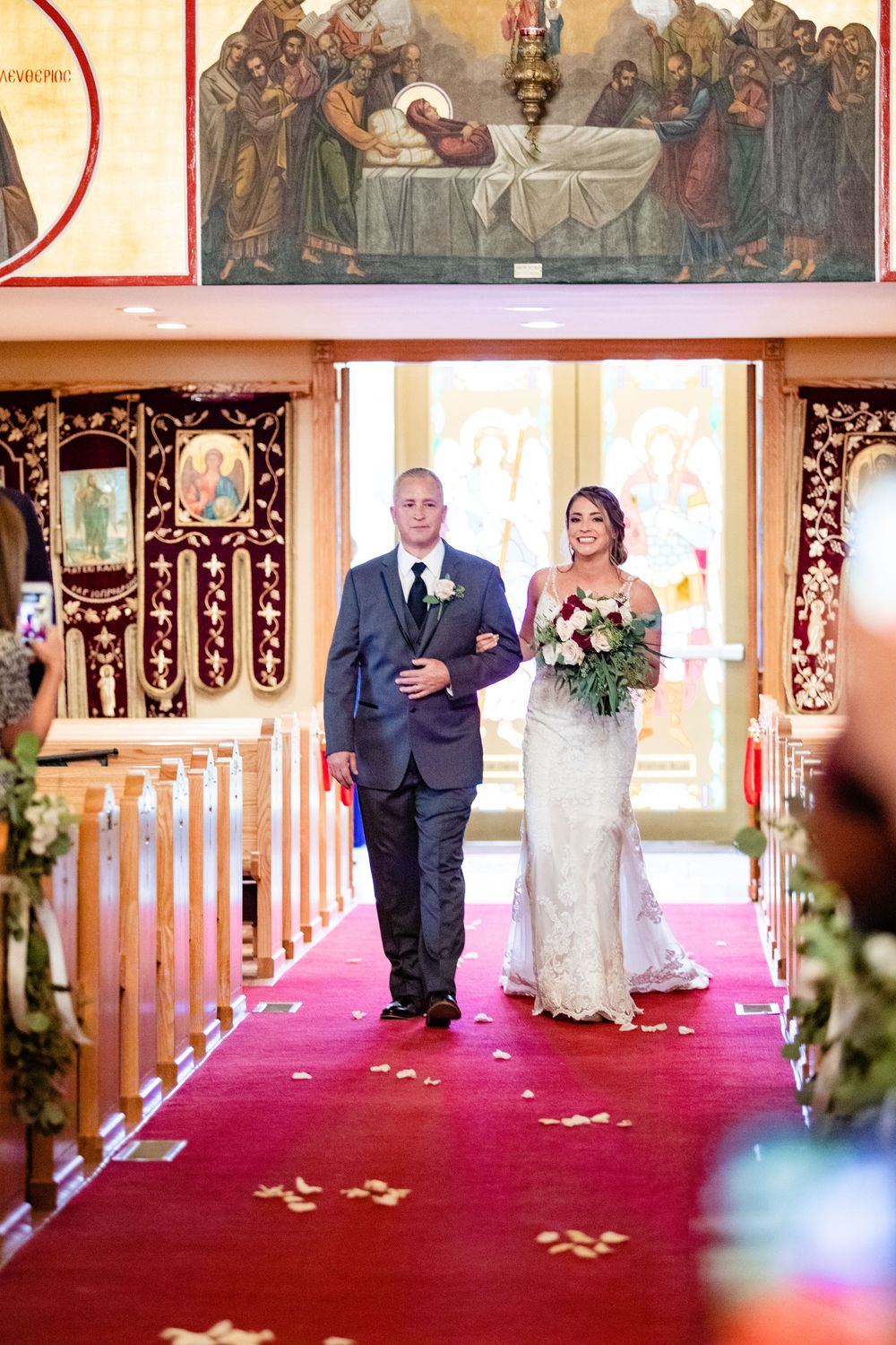 Bride and groom walk down red carpet aisle in ornate Orthodox church adorned with religious icons.