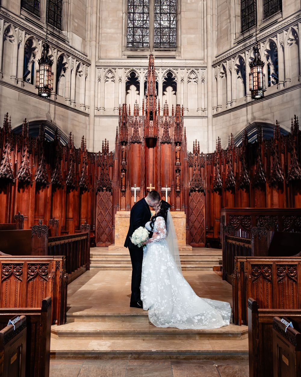 Bride and groom share a kiss at the altar of an ornate Gothic church with wooden pews and intricate carved details.