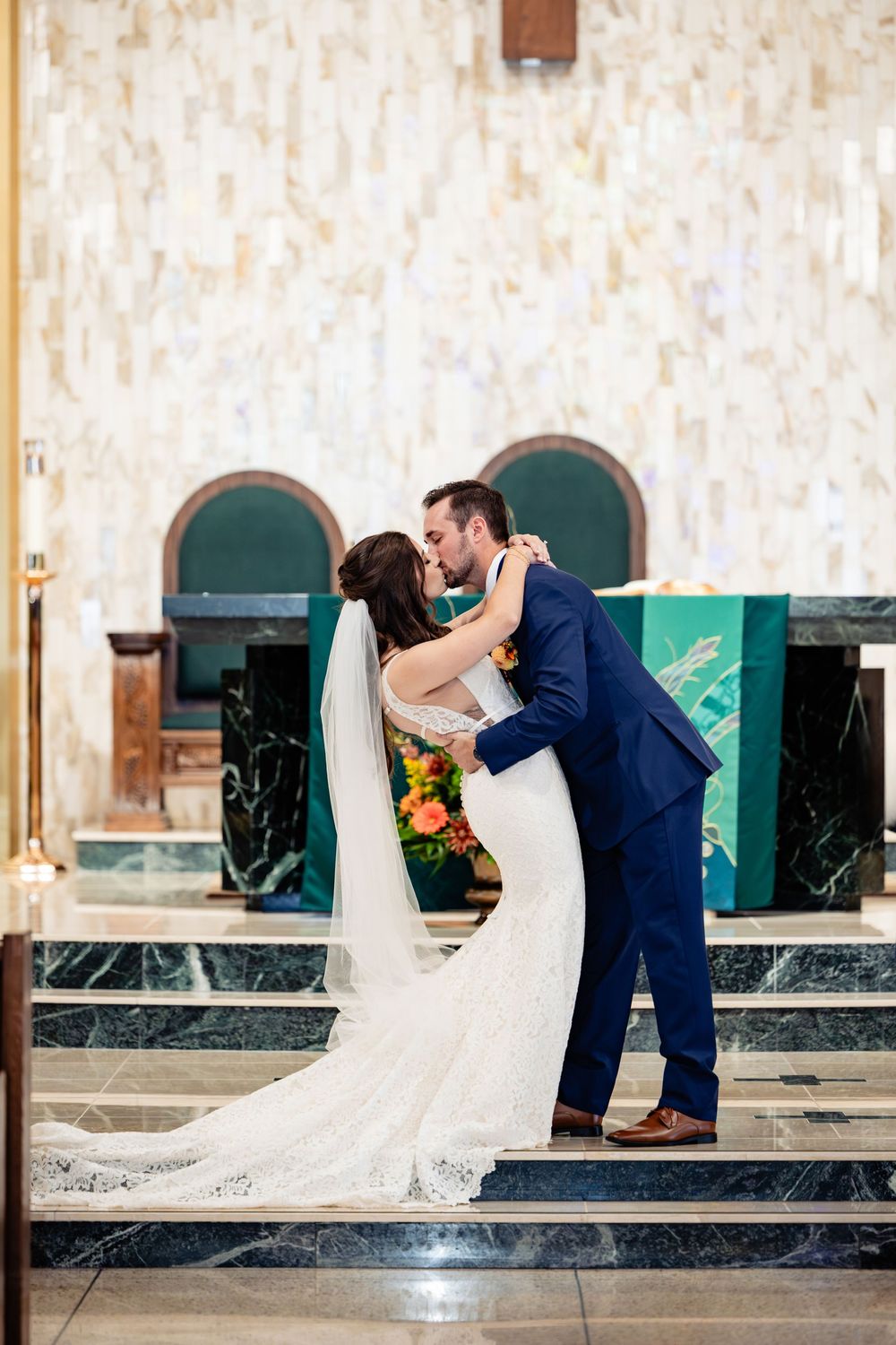 Bride and groom share first kiss at church altar, bride in lace gown with veil, groom in navy suit.