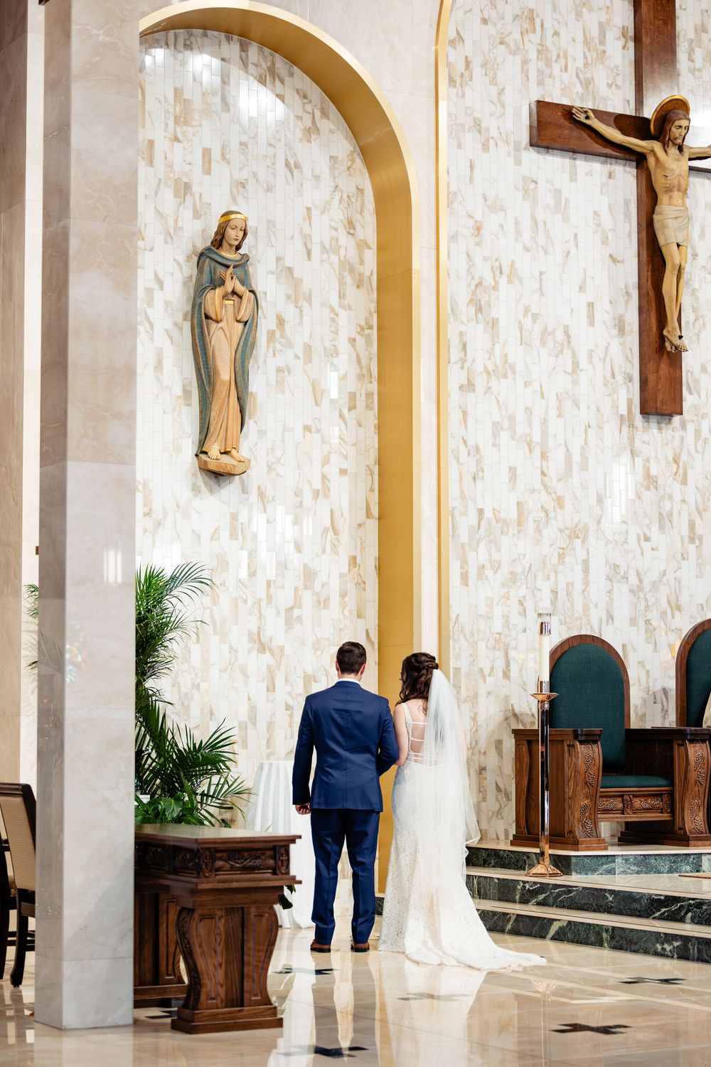 Bride and groom stand before altar in elegant Catholic church with Virgin Mary statue and crucifix.