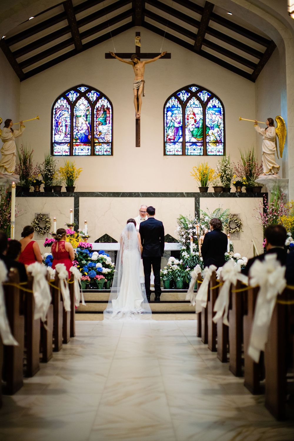 Bride and groom stand at the altar of a Catholic church with stained glass windows and crucifix during wedding ceremony.