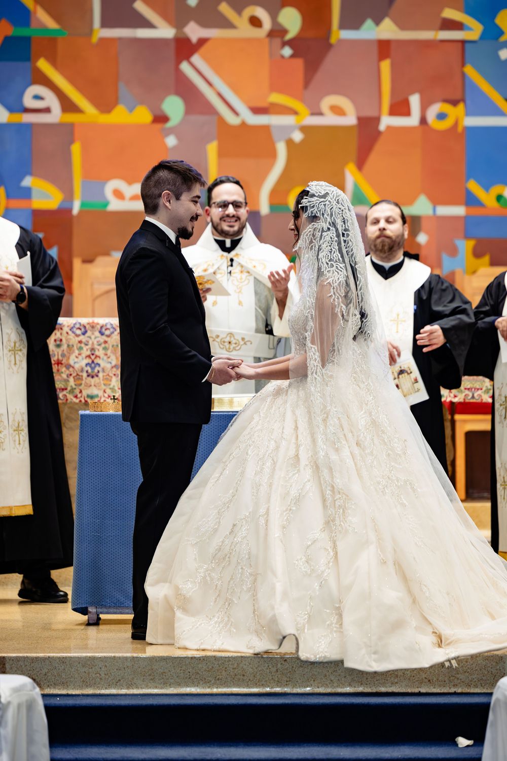 Bride and groom exchange vows at elegant church wedding ceremony with priests officiating before colorful Arabic mural backdrop.