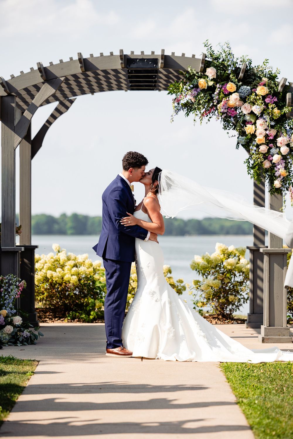 Bride and groom share first kiss under floral pergola arch by lakeside on sunny wedding day.