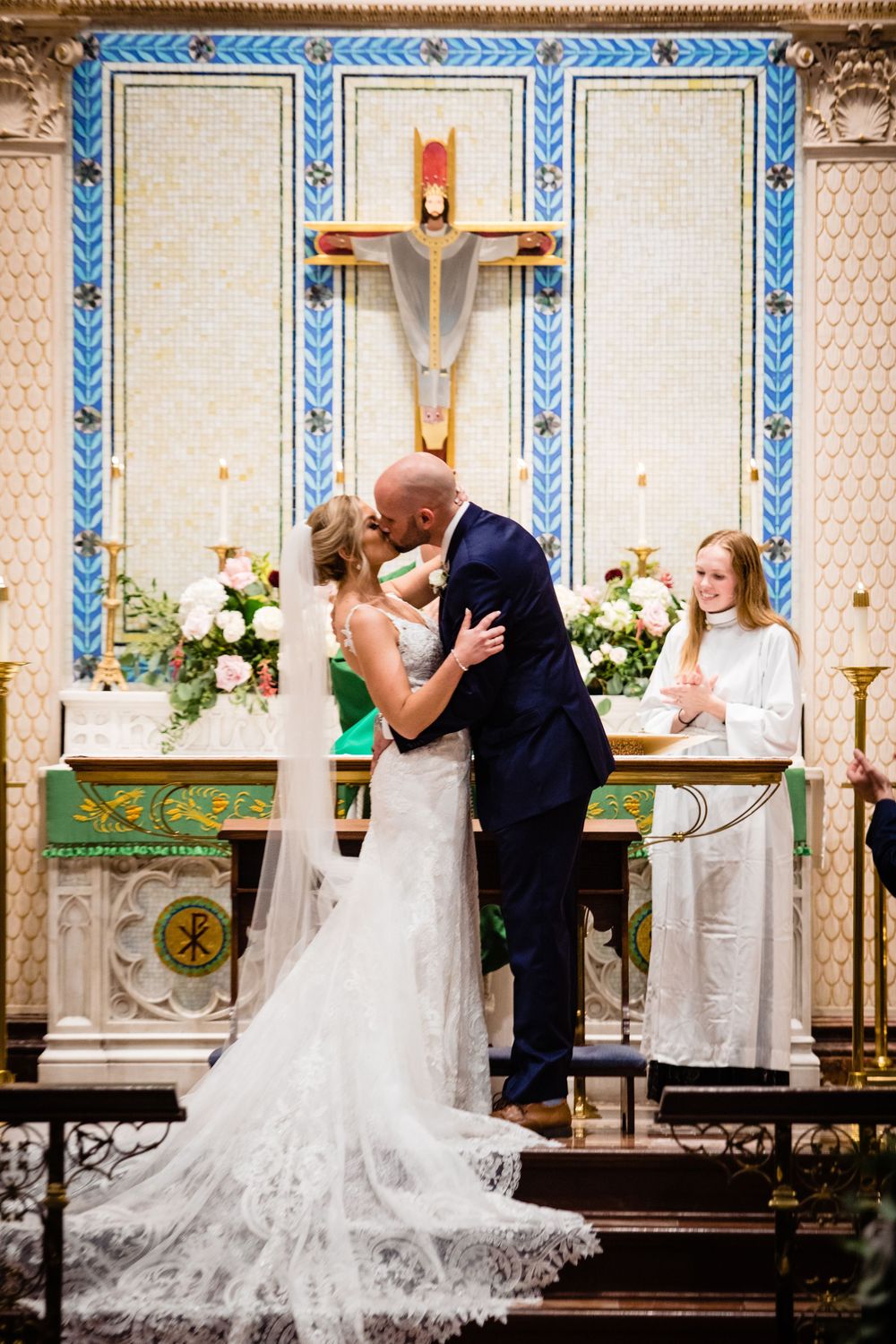 Bride and groom share first kiss at church altar with decorative cross backdrop and flower arrangements.