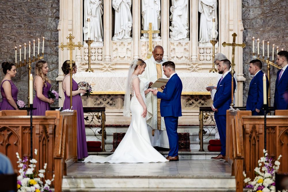 Bride and groom exchange vows at church altar with bridesmaids in purple dresses and groomsmen during wedding ceremony.