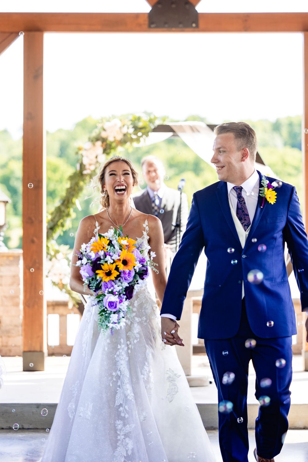 Joyful newlyweds walk hand-in-hand through bubbles, bride holding sunflower bouquet in white gown, groom in blue suit.