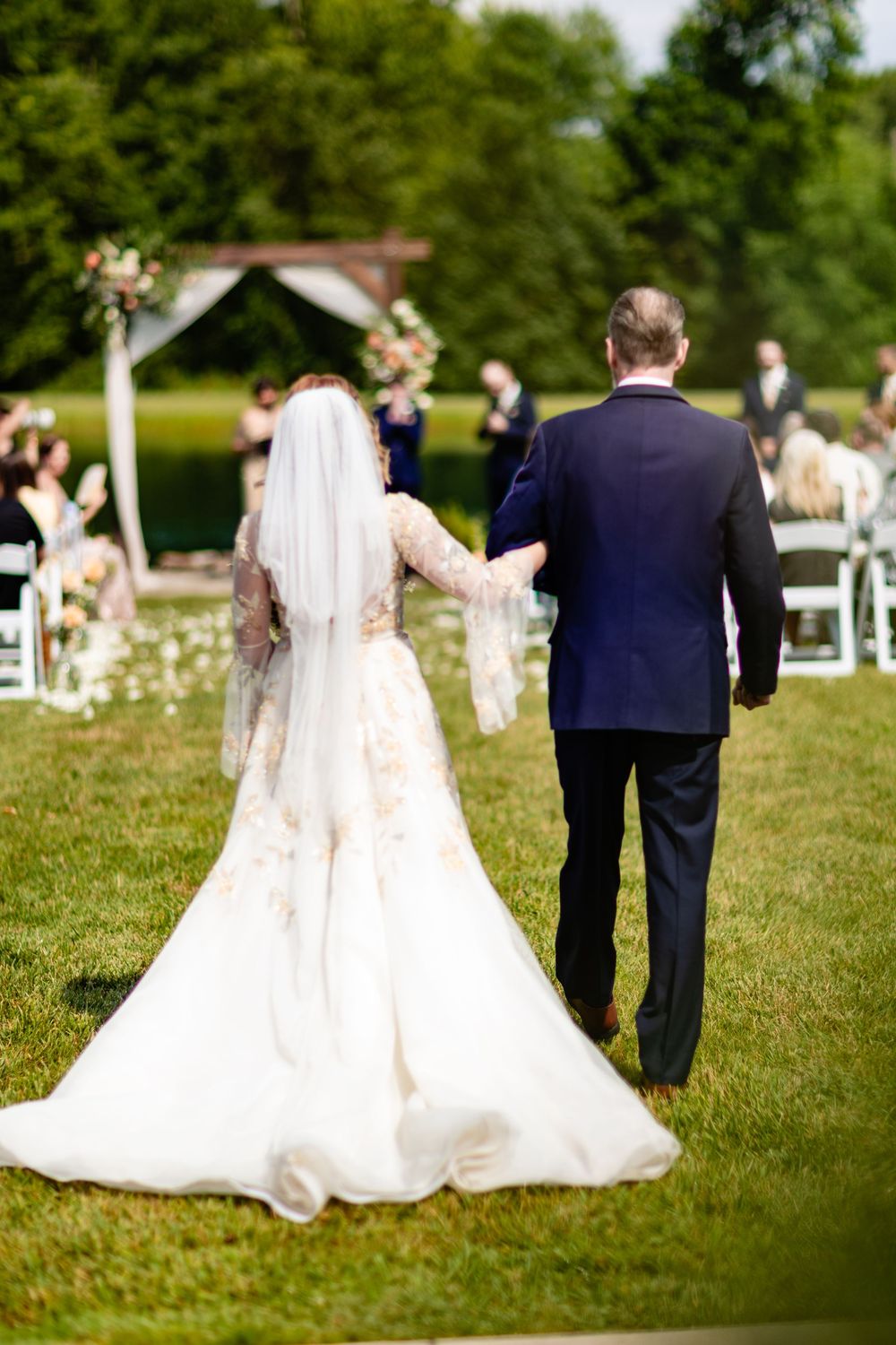 Bride in white gown and veil walks arm-in-arm with groom in navy suit toward outdoor wedding ceremony altar.