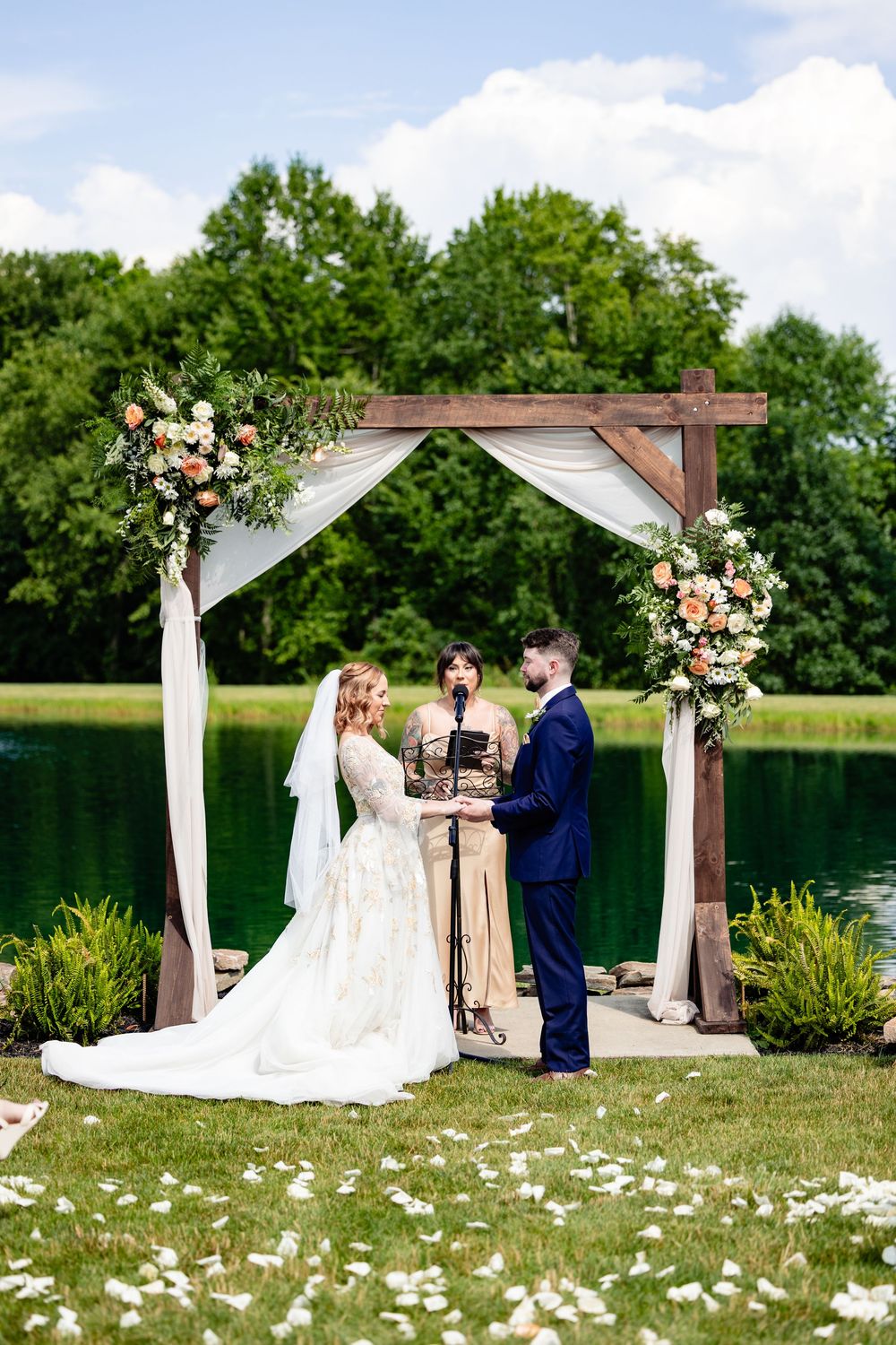 Bride and groom exchange vows under a rustic wooden arch with floral arrangements beside a serene lake.