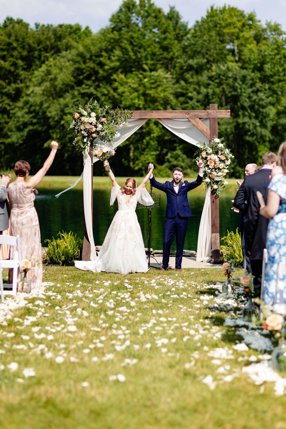 Bride and groom celebrate at outdoor wedding ceremony under floral arch by a lake, surrounded by guests on a sunny day.