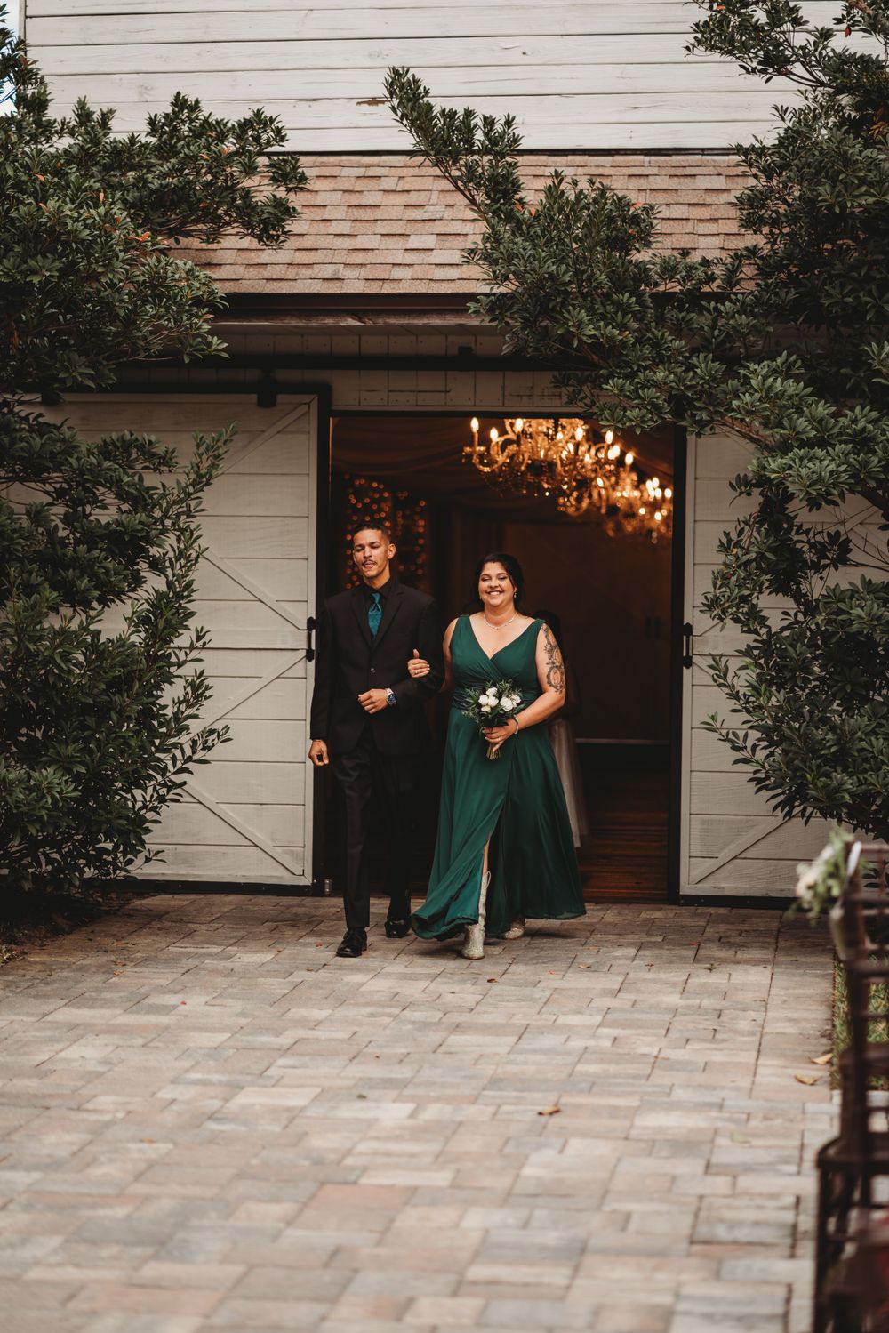 Wedding party members in emerald attire exit rustic barn venue onto stone pathway.