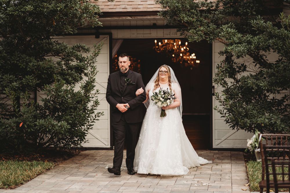 Wedding ceremony processional outside rustic white barn with hanging lights.