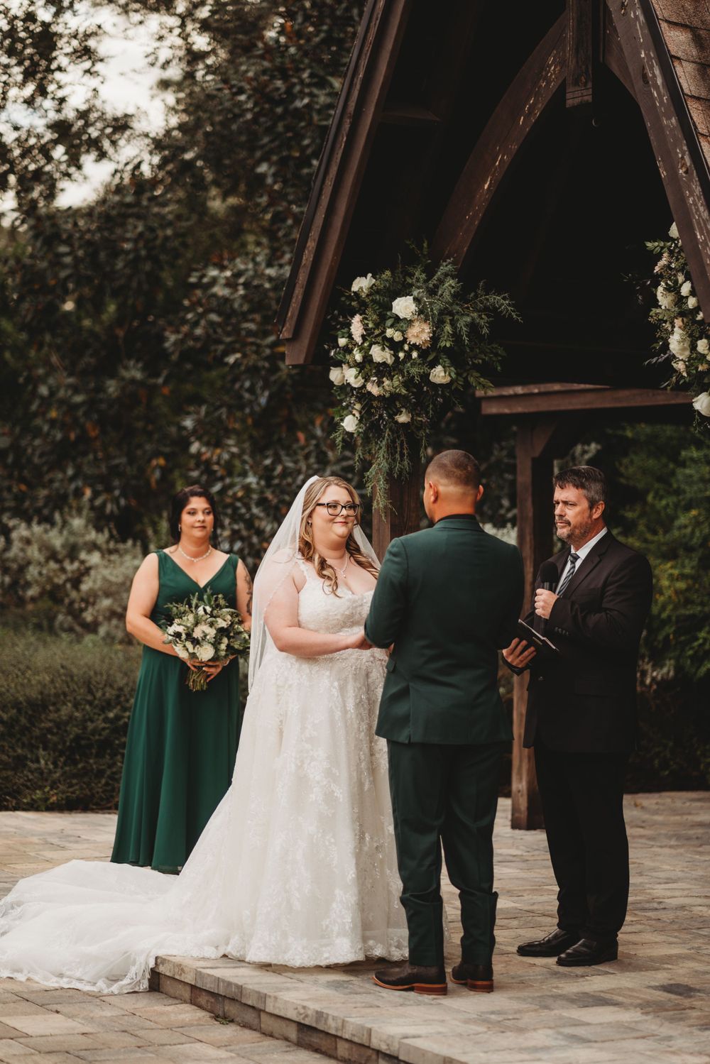 Outdoor wedding ceremony under wooden arch with floral arrangements and emerald bridesmaid dresses.