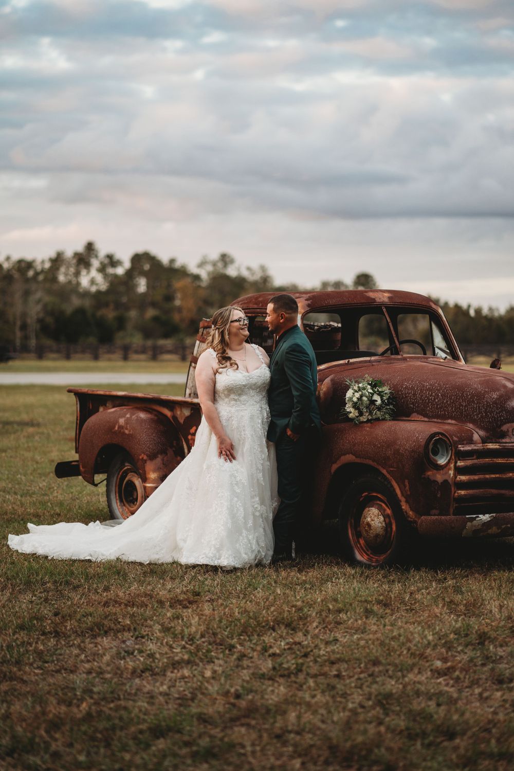 Newlyweds pose romantically against rustic vintage truck in countryside setting.