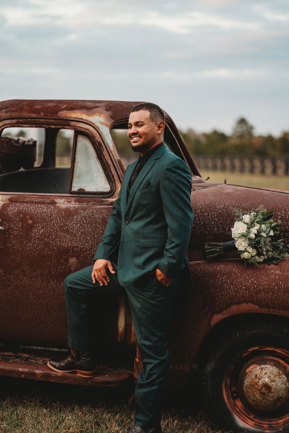 Groom in emerald suit poses beside vintage rustic truck with floral decorations.