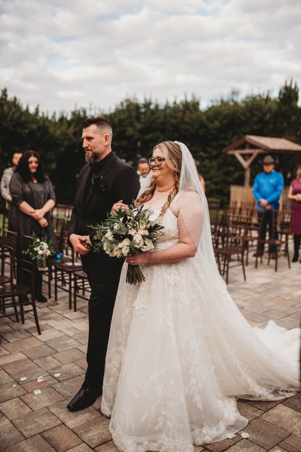 Wedding ceremony moment on stone patio with rustic barn backdrop and wooden chairs.