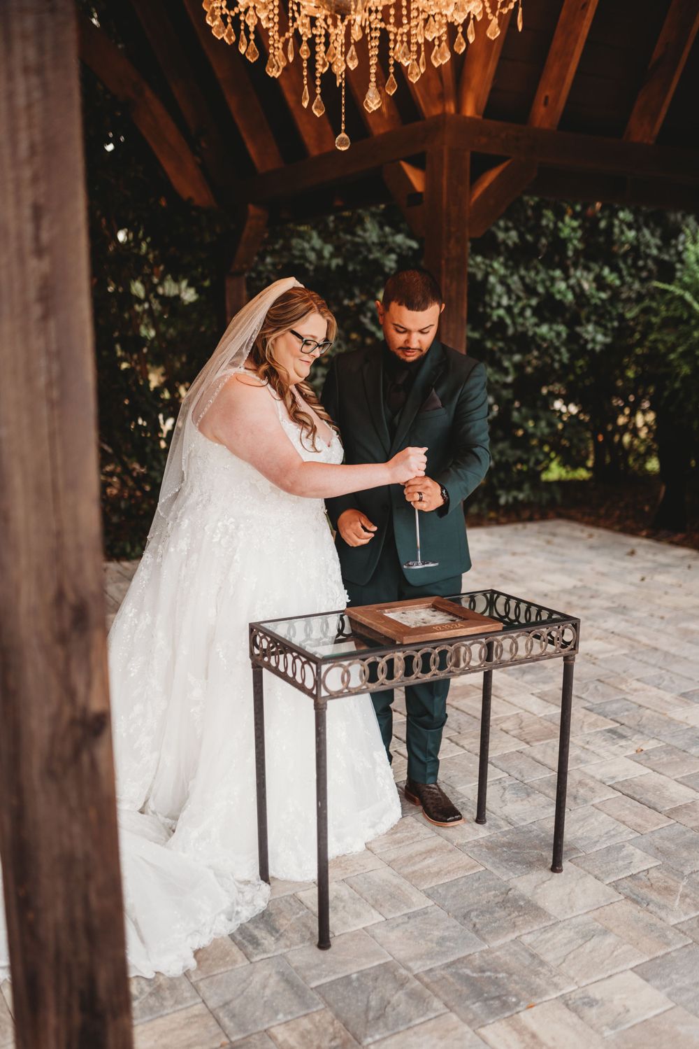 Wedding couple performing unity ceremony at ornate metal table under wooden gazebo with crystal chandelier.