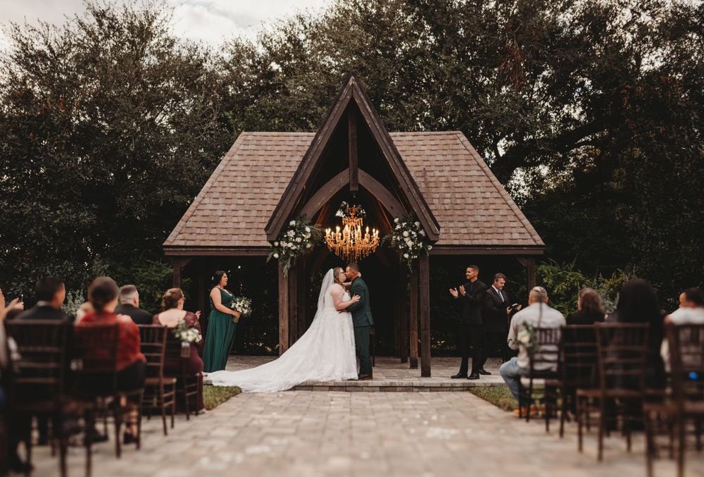 Outdoor wedding ceremony under wooden chapel with chandelier and rows of wooden chairs.