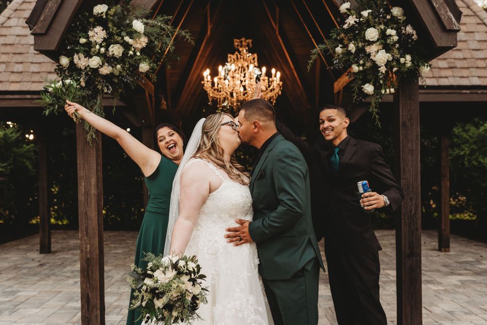 Wedding party celebrating joyfully under wooden chapel with floral arrangements and chandelier.