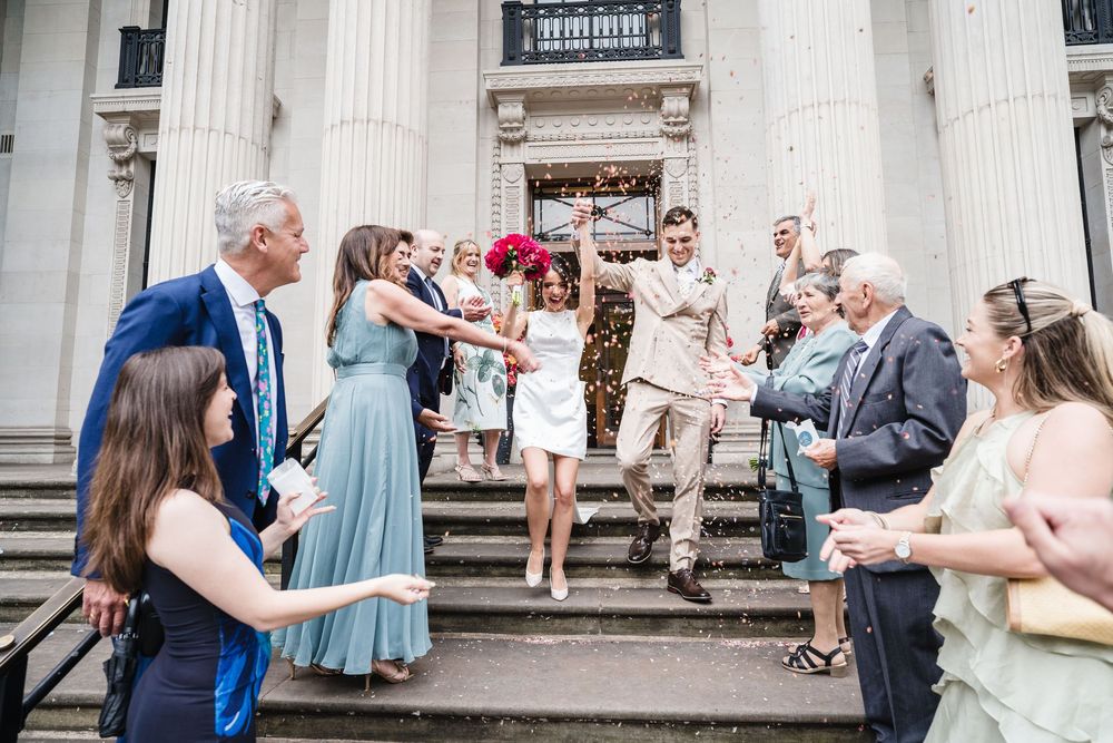 Wedding guests walking single-file along the white columns of a historic building during a sunny outdoor celebration.