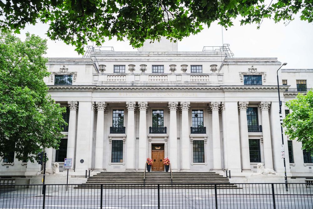 Elegant white neoclassical building with grand columns and ornate architectural details, framed by green tree branches.
