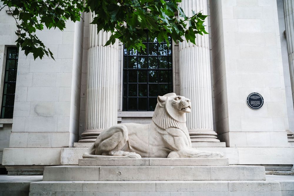 Stone lion statue guards the entrance of a classical building with white columns and a blue historical marker plaque.